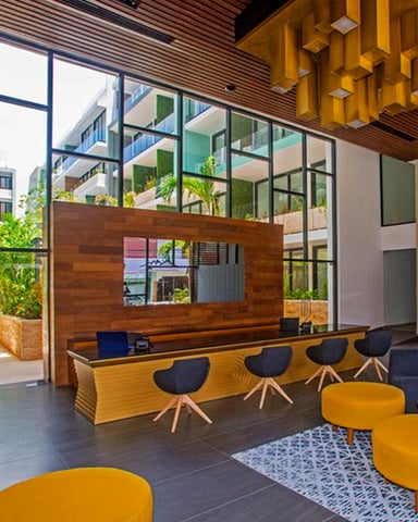Modern lobby with wooden reception desk, yellow pendant light, black chairs, mustard-colored seating, and floor-to-ceiling windows overlooking courtyard with