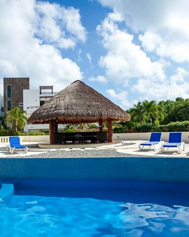 Resort pool with blue loungers, thatched-roof palapa, and modern buildings surrounded by tropical vegetation