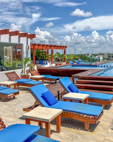 Rooftop pool deck with lounge chairs, blue cushions, wooden pergolas, and infinity pool overlooking city skyline