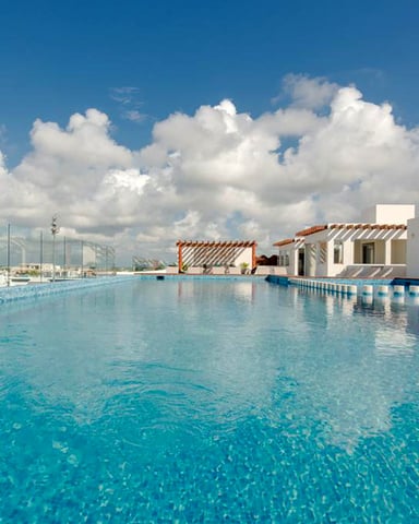 Rooftop infinity pool with turquoise water, white building structures, and city skyline under blue sky with white clouds