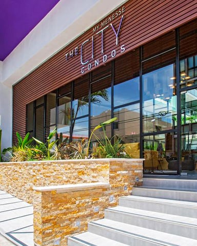 Modern condo building entrance with stone planter boxes, tropical plants, and large glass doors under purple and white awning