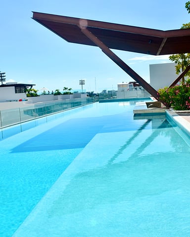 Rooftop swimming pool with clear blue water, dark shade structure, white lounge chairs, and city skyline view