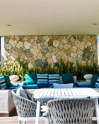 Modern patio with white dining table, woven chairs, built-in seating with teal cushions, stone accent wall, and potted plants