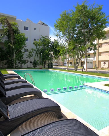 Resort swimming pool with lounge chairs, surrounded by apartment buildings and tall trees under clear blue sky