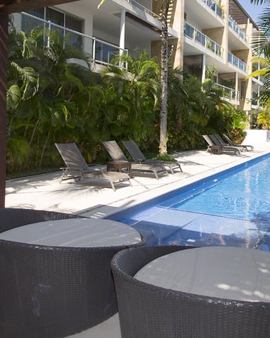 Resort pool with lounge chairs, palm trees, and modern apartment building in background
