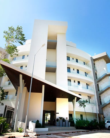 Modern white multi-story apartment building with angular architecture and large covered entrance, surrounded by tropical trees