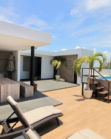Modern waterfront villa with wooden deck, lounge chair, dining area, and infinity pool overlooking turquoise water