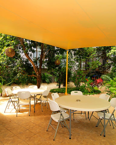Shaded courtyard with round tables, white chairs, and lush tropical plants under yellow awning
