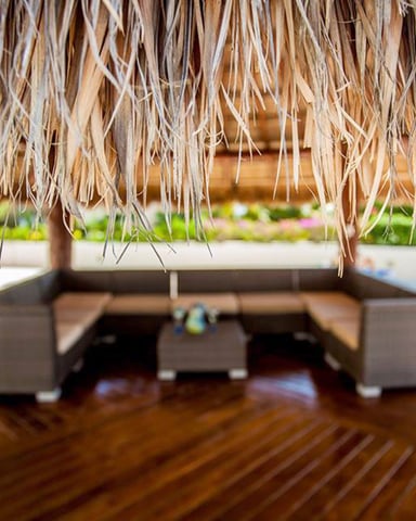 Thatched roof overhang with wooden deck and lounge seating area below, tropical garden visible beyond