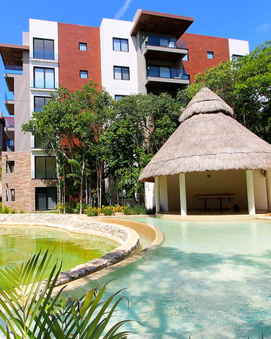 Resort courtyard with turquoise pool, thatched-roof palapa, modern red and white apartment buildings, and tropical vegetation