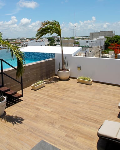 Rooftop terrace with wooden deck, infinity pool, lounge chairs, potted palms, and city skyline view