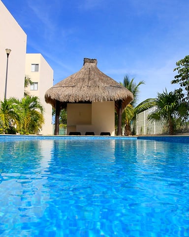Swimming pool with thatched-roof palapa, cream-colored apartment building, and palm trees under blue sky