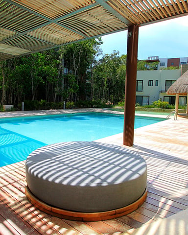 Resort pool area with wooden pergola, round lounge seating, thatched-roof cabana, and modern building in background
