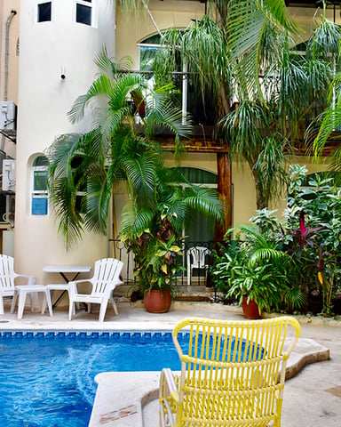 Small courtyard pool with yellow and white lounge chairs, surrounded by lush tropical plants and cream-colored building walls