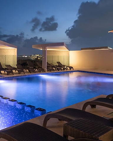 Rooftop pool at dusk with illuminated blue water, lounge chairs, cabanas, and city skyline in background
