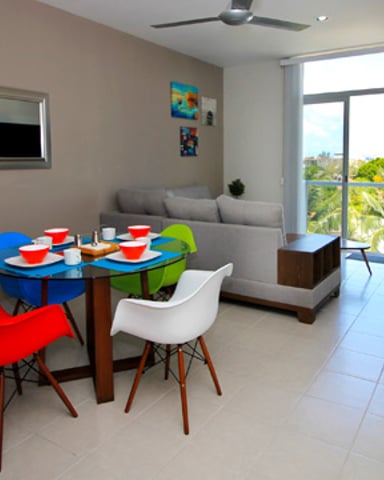 Open-plan living room with colorful dining table, red and white chairs, sofa, and glass doors opening to balcony