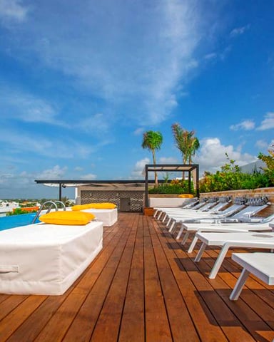 Rooftop pool deck with wooden loungers, blue infinity pool, and tropical plants under clear sky