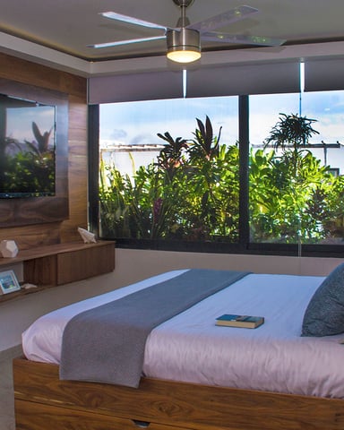 Modern bedroom with wooden platform bed, wall-mounted TV, large windows overlooking tropical plants and ocean view.