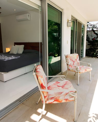 Bedroom with queen bed opening to covered patio with two coral patterned chairs and tropical garden view