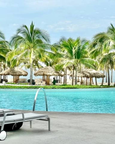 Resort pool with lounge chairs, umbrella, and palm trees with thatched beach cabanas and ocean view