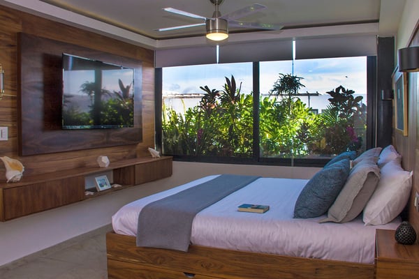 Modern bedroom with wooden platform bed, wall-mounted TV, large windows overlooking tropical plants and ocean view.