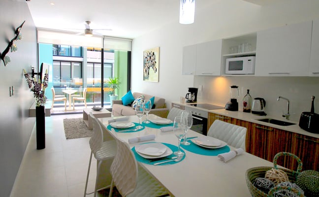 Open-plan kitchen and dining area with turquoise place settings, white chairs, and living room with balcony access beyond